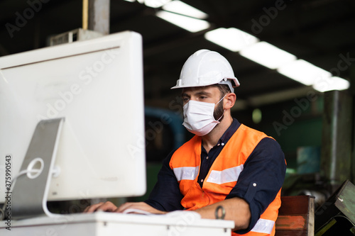 Young caucasian engineering man worker working on digital tablet computer at manufacturing. Worker man wearing face mask prevent covid-19 virus and protective hard hat.