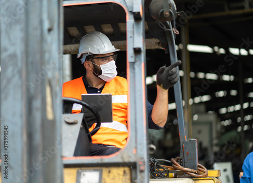 Young male staff driving forklift in warehouse. Worker man wearing face mask prevent covid-19 virus and protective hard hat. Industrial and industrial workers concept