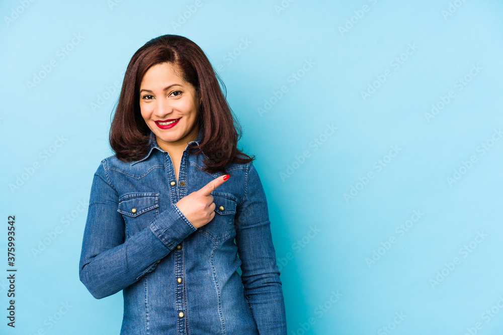 Middle age latin woman isolated on a blue background smiling and pointing aside, showing something at blank space. Middle age latin woman isolated on a blue background smiling and pointing aside, showing something at blank space.