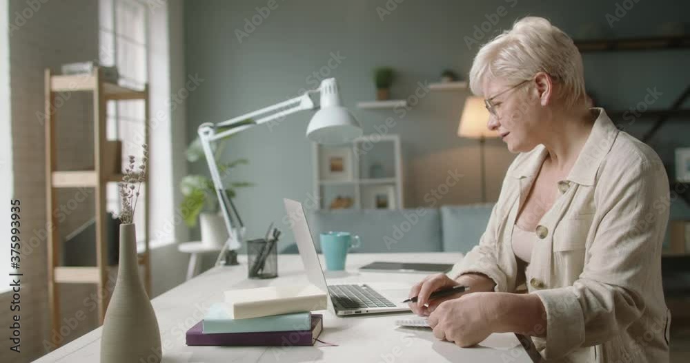 Senior caucasian lady working on laptop. Mature woman in 50s using her computer at home, typing on keyboard, surfing web and making notes 4k footage