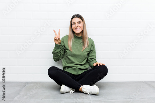 Young caucasian woman sitting on the floor showing victory sign and smiling broadly.