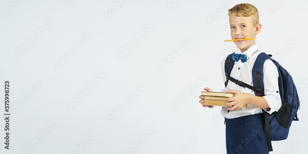 A schoolboy with a briefcase holds books and a pencil in his teeth. Looks at them. Isolated background. A schoolboy with a briefcase holds books and a pencil in his teeth. Looks at them. Isolated background.