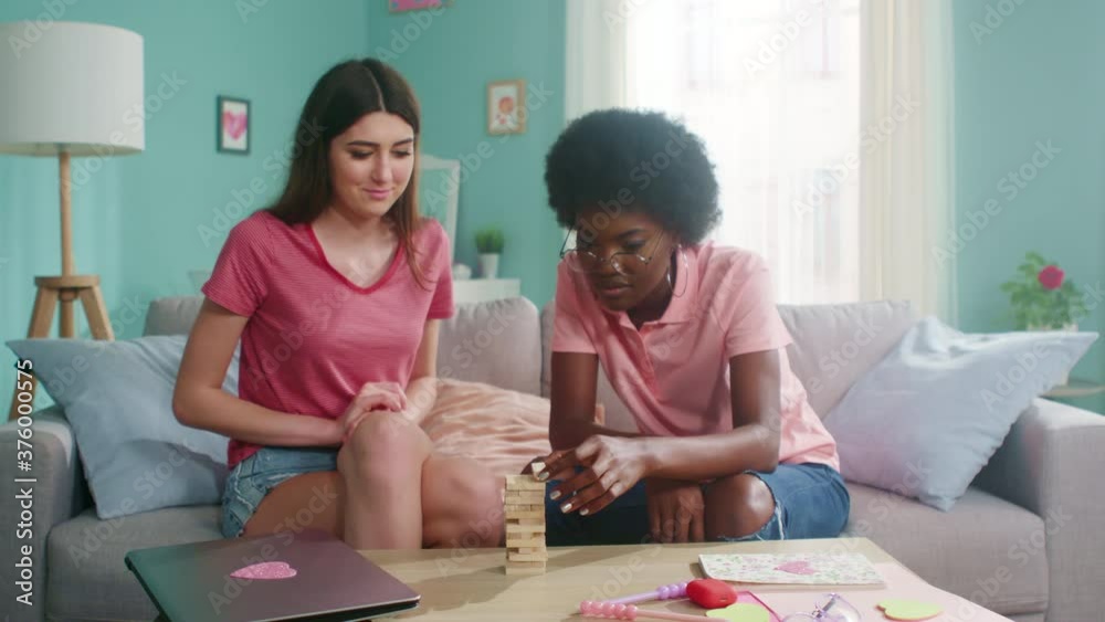 Young women, roommates, have fun playing interesting board game, constructing wooden tower of small bars, enjoy home leisure, Foreground, Slow motion. Young women, roommates, have fun playing interesting board game, constructing wooden tower of small bars, enjoy home leisure, Foreground, Slow motion.