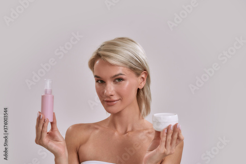 Portrait of young female blonde with perfect fresh skin looking at camera, holding jar and bottle of different cosmetic skincare products while posing isolated over grey background