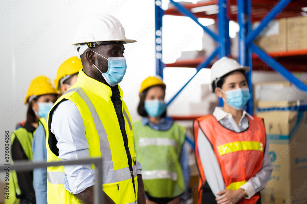 Obraz premium Warehouse managers and worker wearing face mask prevent covid-19 virus and protective hard hat and working together in warehouse workplace. Professional engineering team.