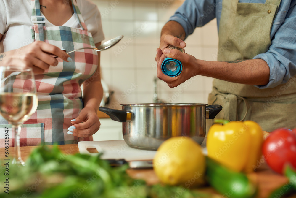 Cropped shot of man adding pepper, spice to the soup while woman holding a spoon. Couple preparing a meal together in the kitchen. Cooking at home, Italian cuisine