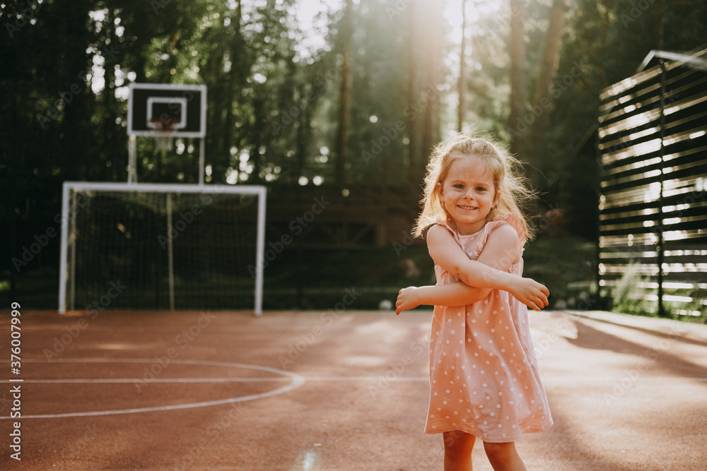 Naklejka premium Cute little girl in pink casual dress smiling standing on sports court at sunset.