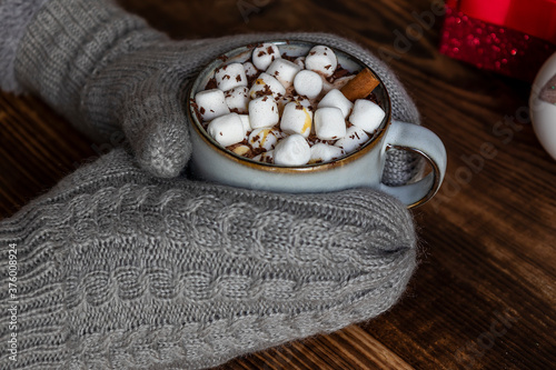 Cozy holiday composition, hands in mittens keep mug with cocoa or chocolate with marshmallow.