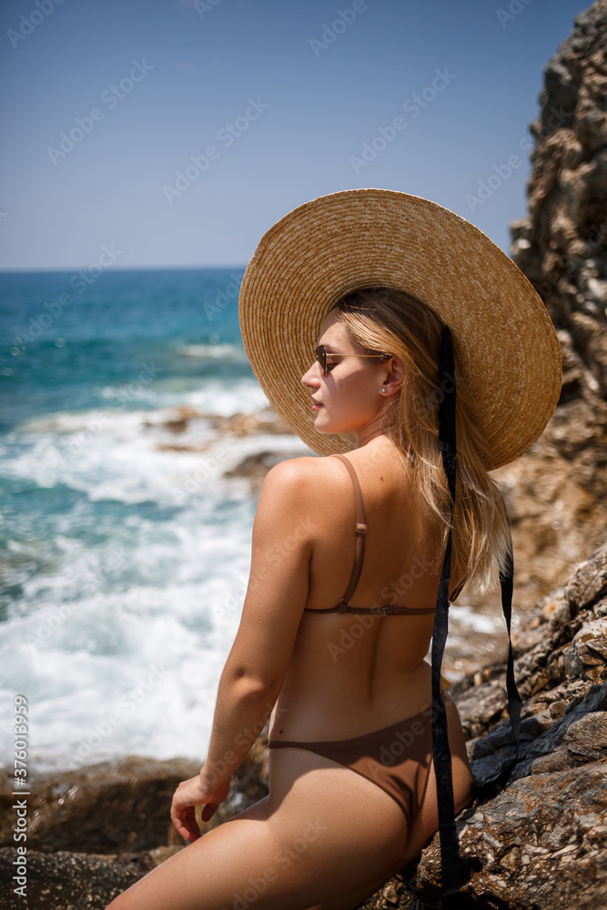 A beautiful girl in a swimsuit with a golden tan in a hat stands near the rocky coast. Summer sunny day by the sea. Vacation concept