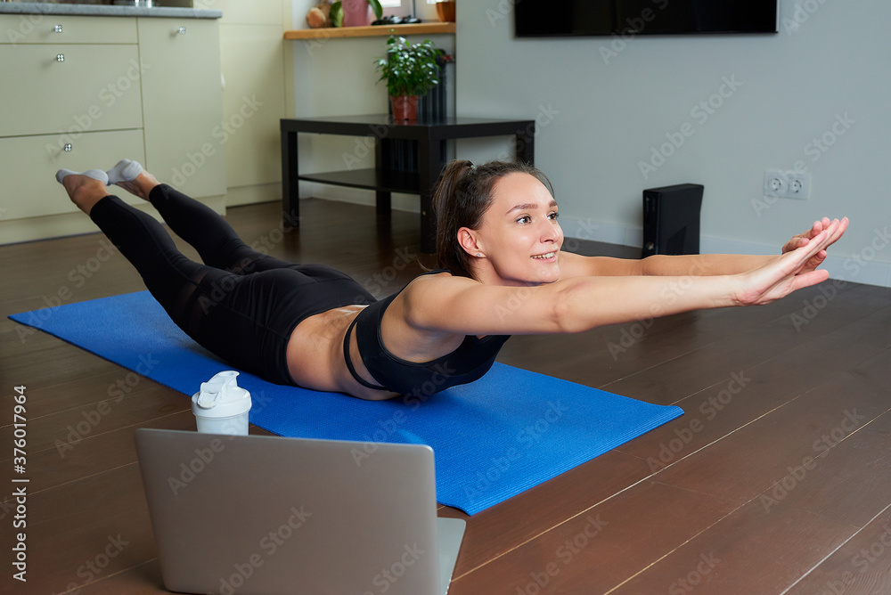 A smiling sporty girl in a black workout tight suit is doing the classical boat exercise for the back. A coach in a superman pose conducting a remote online fitness class on the blue yoga mat at home.