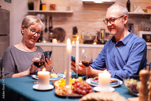 Pensioner man smiling using phone and having a conversation with wife in kitchen in time of dinner. Sitting at the table in the dining room  browsing  searching  using phone  internet 