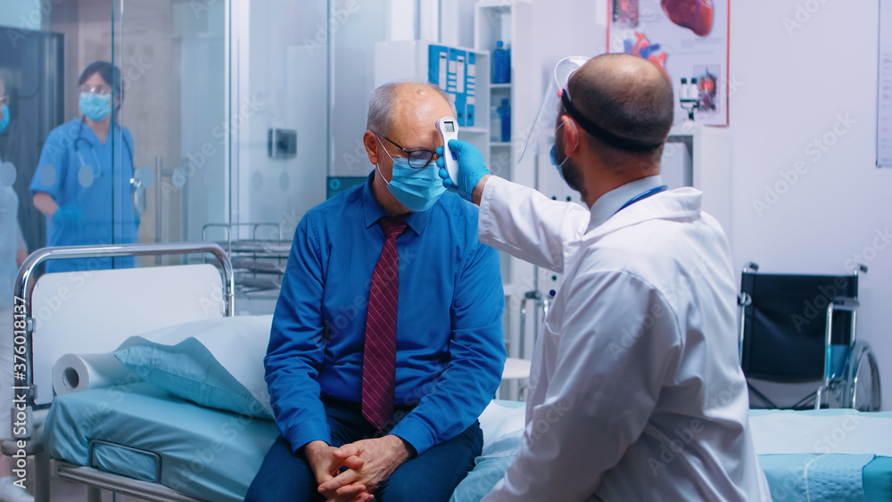 Doctor checking old man temperature with a infrared thermometer. Retired senior man wearing a mask and healthcare worker in protective equipment for consultation. Modern private clinic Doctor checking old man temperature with a infrared thermometer. Retired senior man wearing a mask and healthcare worker in protective equipment for consultation. Modern private clinic