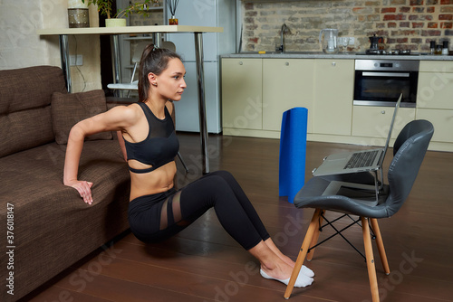 Obraz na płótnie A sporty girl in a black tight suit is doing triceps and chest workouts and watching an online training video on a laptop. A female coach conducting a remote fitness class at home. w salonie