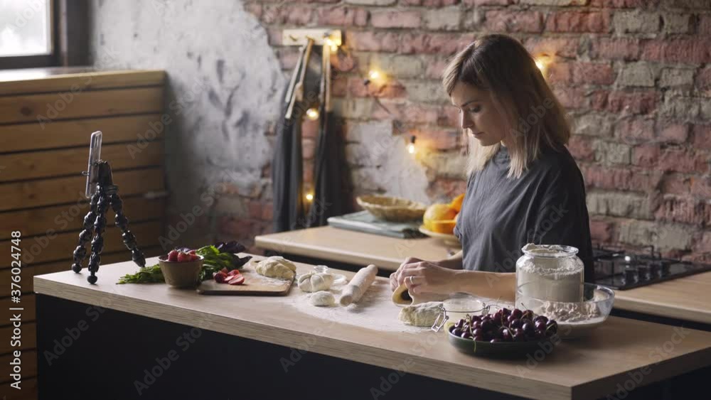 young woman with fair hair prepares cherry cake standing at brown wooden kitchen table with fresh products and smartphone on tripod at home young woman with fair hair prepares cherry cake standing at brown wooden kitchen table with fresh products and smartphone on tripod at home