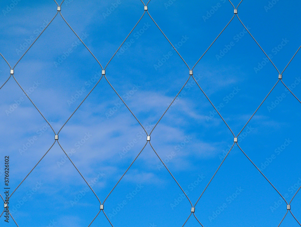 Abstract view of stainless steel mesh fence with clamps and clear blue sky background with light soft white clouds. flat on view. summer scene. modern construction materials concept. fine wire cable. Abstract view of stainless steel mesh fence with clamps and clear blue sky background with light soft white clouds. flat on view. summer scene. modern construction materials concept. fine wire cable.