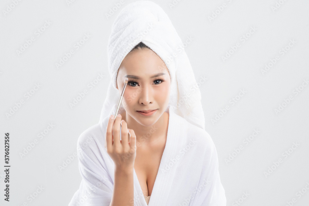 Beautiful young woman wearing bathrobe with towel with towel on head is using a makeup brush makeup her after bathing, at studio shot isolated on white background. Beautiful young woman wearing bathrobe with towel with towel on head is using a makeup brush makeup her after bathing, at studio shot isolated on white background.