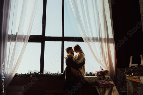 Silhouette of a loving couple, man and woman against of a huge stained glass window with a view of the blue sky and curtains on the sides. The comfort of home, a life together for couple.