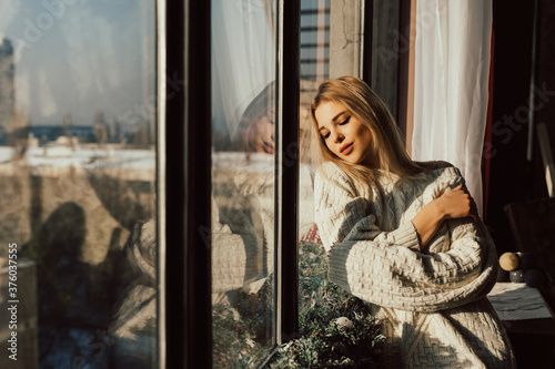 Young attractive woman relaxing on windowsill with eyes closed. Gorgeous blonde young woman in casual clothes having relaxed and enjoying warm sunlight.