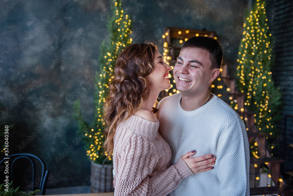 Extra close-up portrait of a happy couple in love on the background of a Christmas tree and garlands of lights. Beautiful girl kisses on the cheek of a young man, lovers laugh, hugs in warm sweater