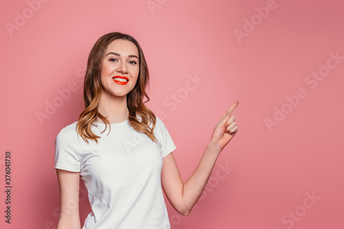 Cheerful smiling girl in white t-shirt with red lipstick smiles, looks at camera and points finger at copy space isolated over pink background