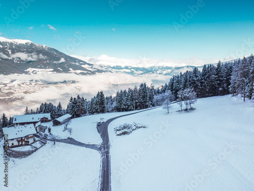 Aerial view of snowy cloudy mountains, forest with a road, small chalet house. Captured from above with a drone. Dolomites Alps- Italy