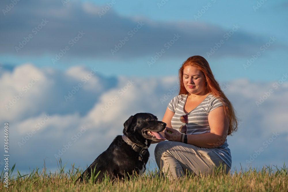 Plump young red-haired blue-eyed girl with a labrador retriever photographed on a field against a background of clouds. Plump young red-haired blue-eyed girl with a labrador retriever photographed on a field against a background of clouds.