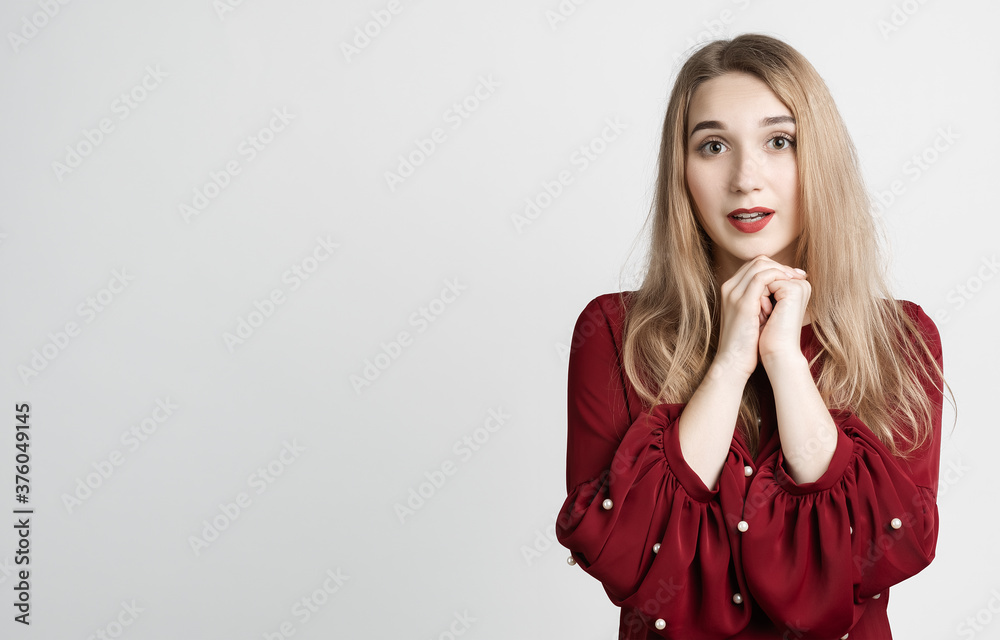 Young woman with long blonde hair keeps hands together near chin, has cute expression, wears red blouse. Copy space Young woman with long blonde hair keeps hands together near chin, has cute expression, wears red blouse. Copy space