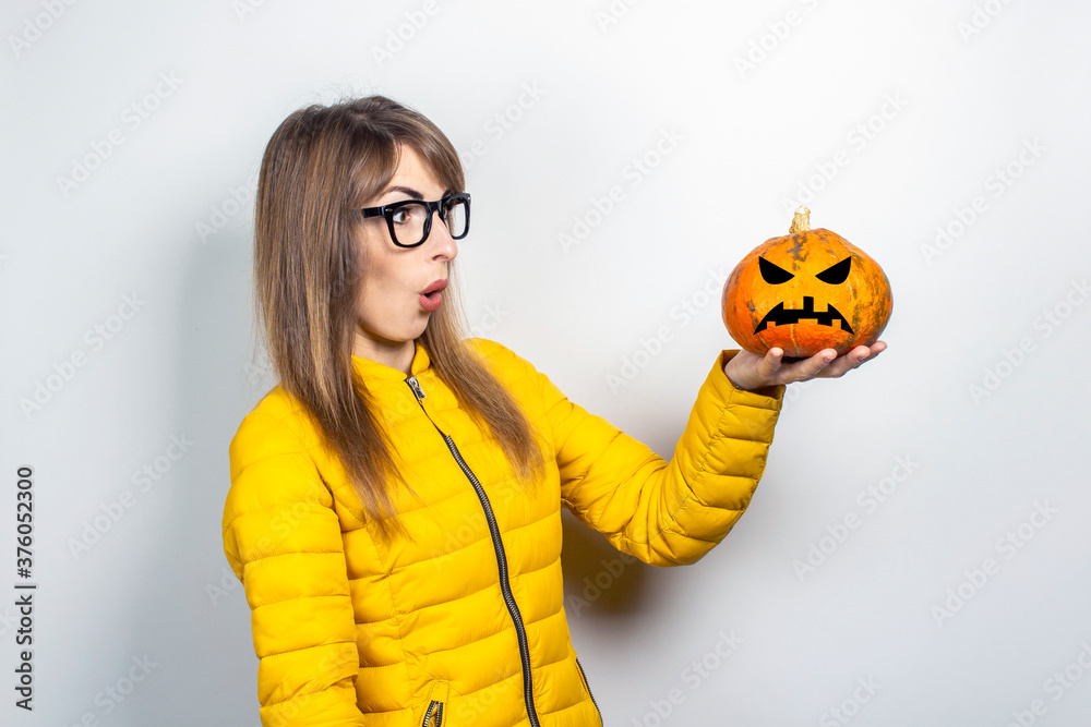 young girl in a yellow jacket holds a pumpkin with a frightened face on a light background. Halloween concept, autumn, celebration young girl in a yellow jacket holds a pumpkin with a frightened face on a light background. Halloween concept, autumn, celebration
