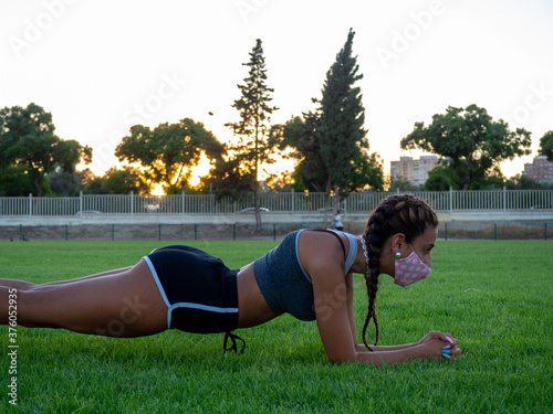 Mujer joven con mascarilla haciendo el ejercicio de plancha en el césped para cumplir las medidas de seguridad y distanciamiento de la pandemia del coronavirus