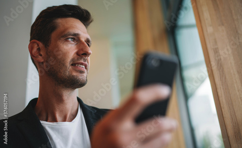 Successful male executive ceo standing at office using smartphone device communicates with colleague, Pensive businessman in casual suit texting on cellular standing near big window in business center