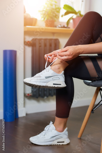 A close photo of the hands and legs of a sporty girl in black tight suit who is tying her shoelaces on white sneakers while sitting on the chair before training at home.