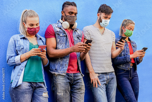 Group of people using their smartphones in covid 19 times protected with face mask - Friends checking online news while standing by the wall and holding cellphones