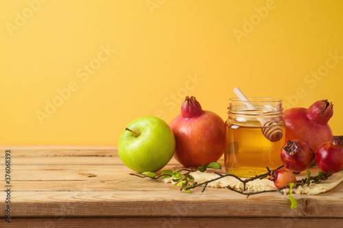 Jewish holiday Rosh Hashana concept with honey jar, apple and pomegranate on wooden table over yellow background
