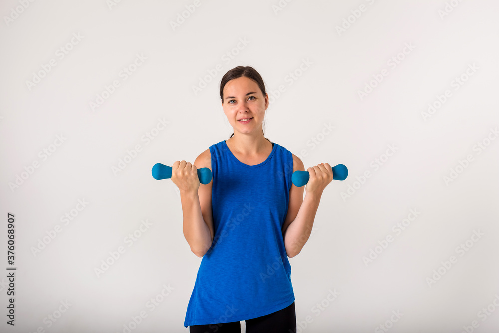 A woman performs exercises with dumbbells on her biceps and looks at the camera on a white background. A woman performs exercises with dumbbells on her biceps and looks at the camera on a white background.