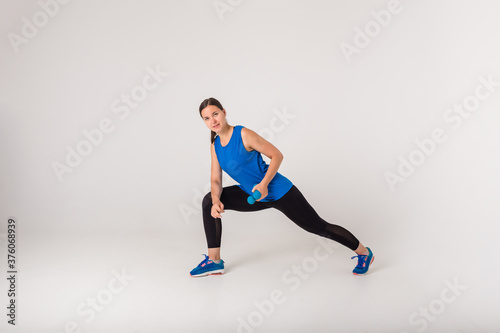 A woman performs a lunge exercise with dumbbells and looks at the camera on a white background.