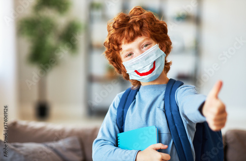 Little ginger schoolboy in protective mask with drawn smile shows thumb up.