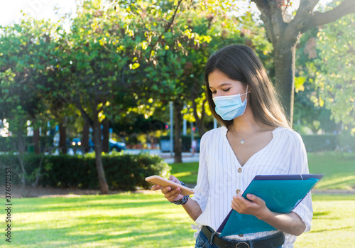 Young woman wearing a medical mask using her phone at the park.