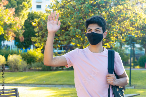 A young man with a medical mask waving to someone in the park.