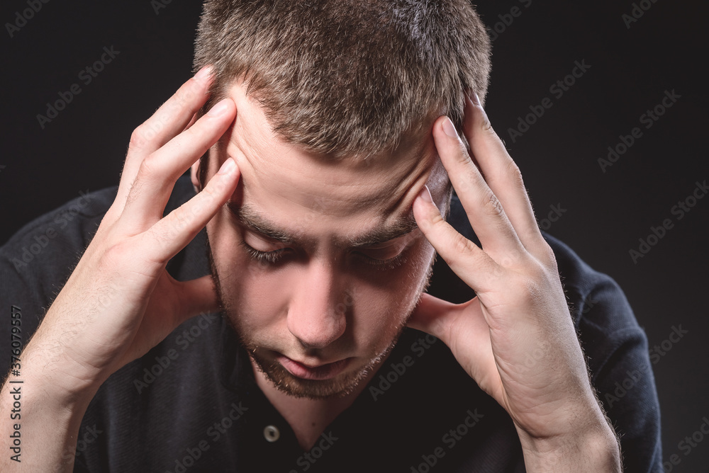 Fototapeta premium Young handsome man in a black shirt sits and holds his head on a black background. Experiencing stress, pain