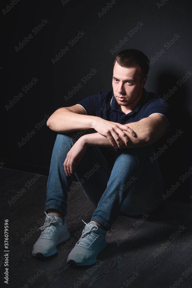 Young attractive man in blue jeans and a polo shirt sits on the floor near the wall in the studio. Youth and attractiveness concept Young attractive man in blue jeans and a polo shirt sits on the floor near the wall in the studio. Youth and attractiveness concept