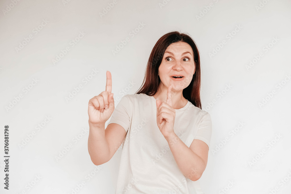 Emotional brunette woman with surprised face keeps holds hands in front of her white t shirt, has minimal makeup, poses indoor on a white background. Copy, empty space for text Emotional brunette woman with surprised face keeps holds hands in front of her white t shirt, has minimal makeup, poses indoor on a white background. Copy, empty space for text