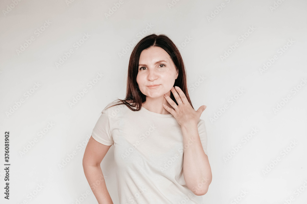 Beautiful brunette woman keeps hands on cheek, dressed in casual white t shirt, has minimal makeup, poses indoor on a white background. Copy, empty space for text Beautiful brunette woman keeps hands on cheek, dressed in casual white t shirt, has minimal makeup, poses indoor on a white background. Copy, empty space for text