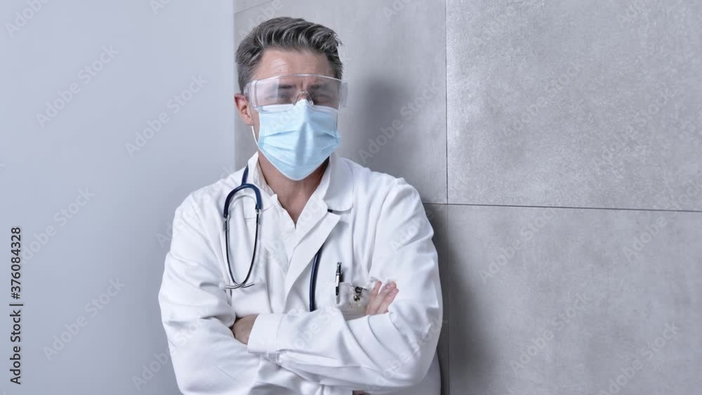 Covid-19 - Portrait of older male doctor in medical virus mask and protective glasses. Man wearing white medical lab coat standing against gray wall. Copy space. Covid-19 - Portrait of older male doctor in medical virus mask and protective glasses. Man wearing white medical lab coat standing against gray wall. Copy space.