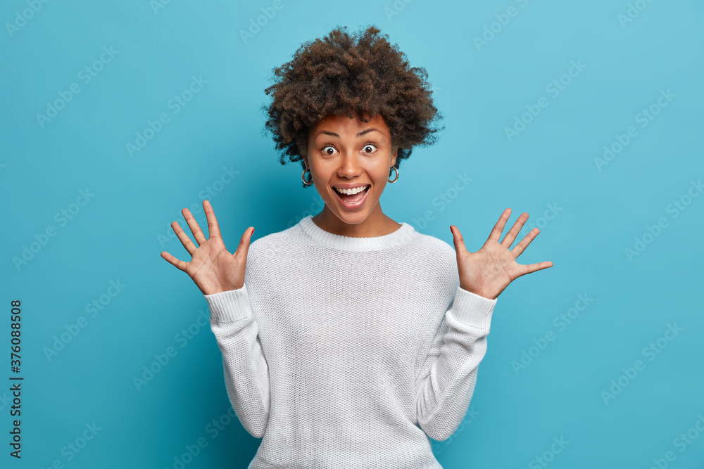 Amused pretty girl with curly Afro hair, raises palms, has cheerful expression, smiles broadly, sees something funny, wears white sweater, isolated on blue background. Happy emotions concept - obrazy, fototapety, plakaty 