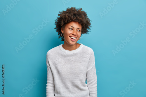 Joyful dark skinned young woman with broad smiles, perfect teeth, feels carefree and enthusiastic, looks happily aside, stands entertained, wears casual white jumper, isolated on blue background