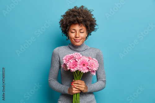 Pleased romantic Afro American woman holds beautiful bouquet of gerbera flowers, has date with boyfriend, closes eyes, wears grey turtleneck, isolated on blue bakground. Womens day, relationships