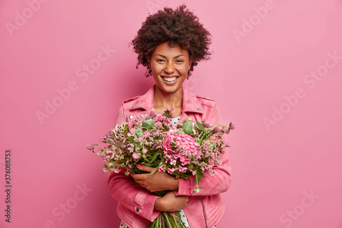 Horizontal shot of pretty African American woman expresses sincere emotions, embraces bouquet of flowers, has spring mood, poses against pink background, gets congratulation on her anniversary