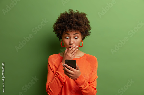 Horizontal shot of amazed adult woman being deeply surprised, stares at smartphone display, reads shocking news on website, dressed in orange jumper, has bugged eyes, covers mouth. Omg, its horrible!