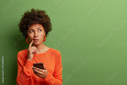 Photo of pretty ethnic woman ponders on how to answer question, thinks deeply about something, uses modern mobile phone, tries to made up good message, keeps index finger near lips, stands indoor photo