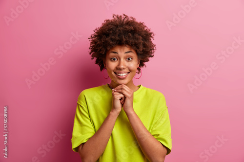 Pretty good looking girl with curly hair, keeps hands under chin, smiles gladfully, has white teeth, wears casual green t shirt, poses against pink studio background, has relaxed face expression